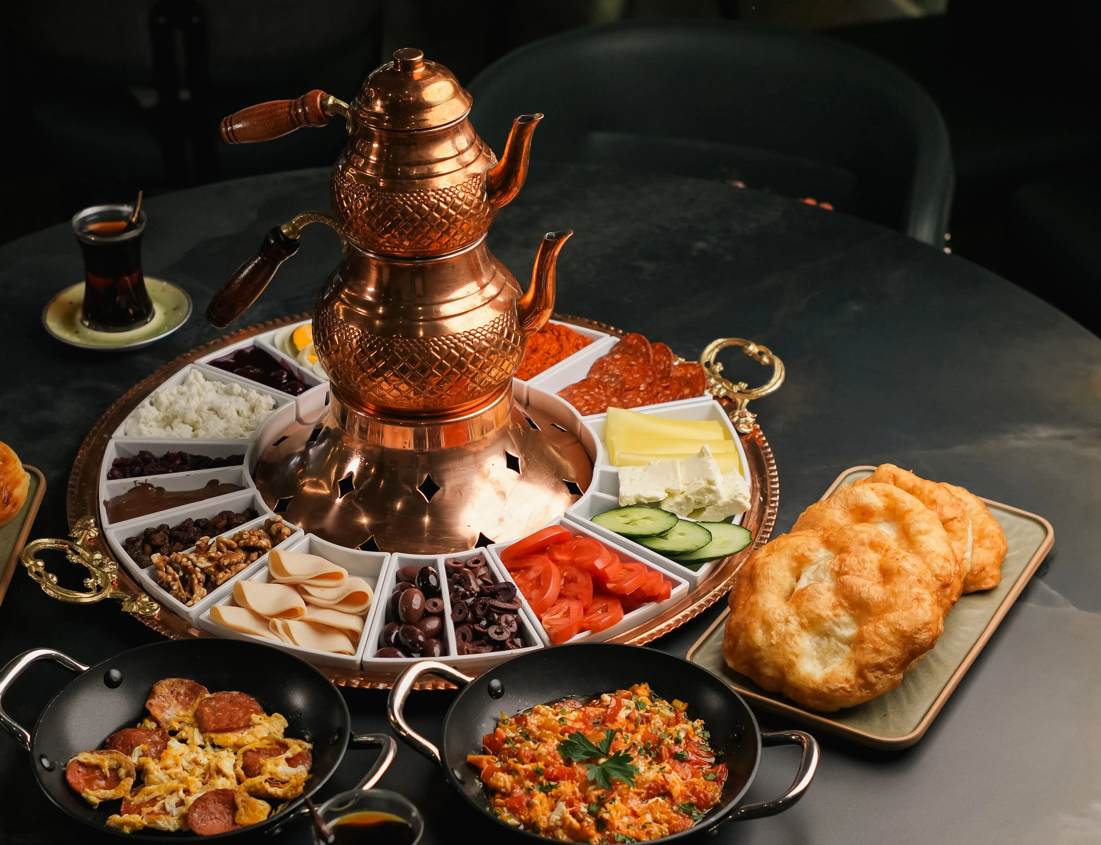 Breakfast for 4: a copper double teapot on a round tray of olives, cheeses, tomatoes, cucumber, walnuts and jam, with pans of menemen and sucuk with egg and a plate of warm fried dough