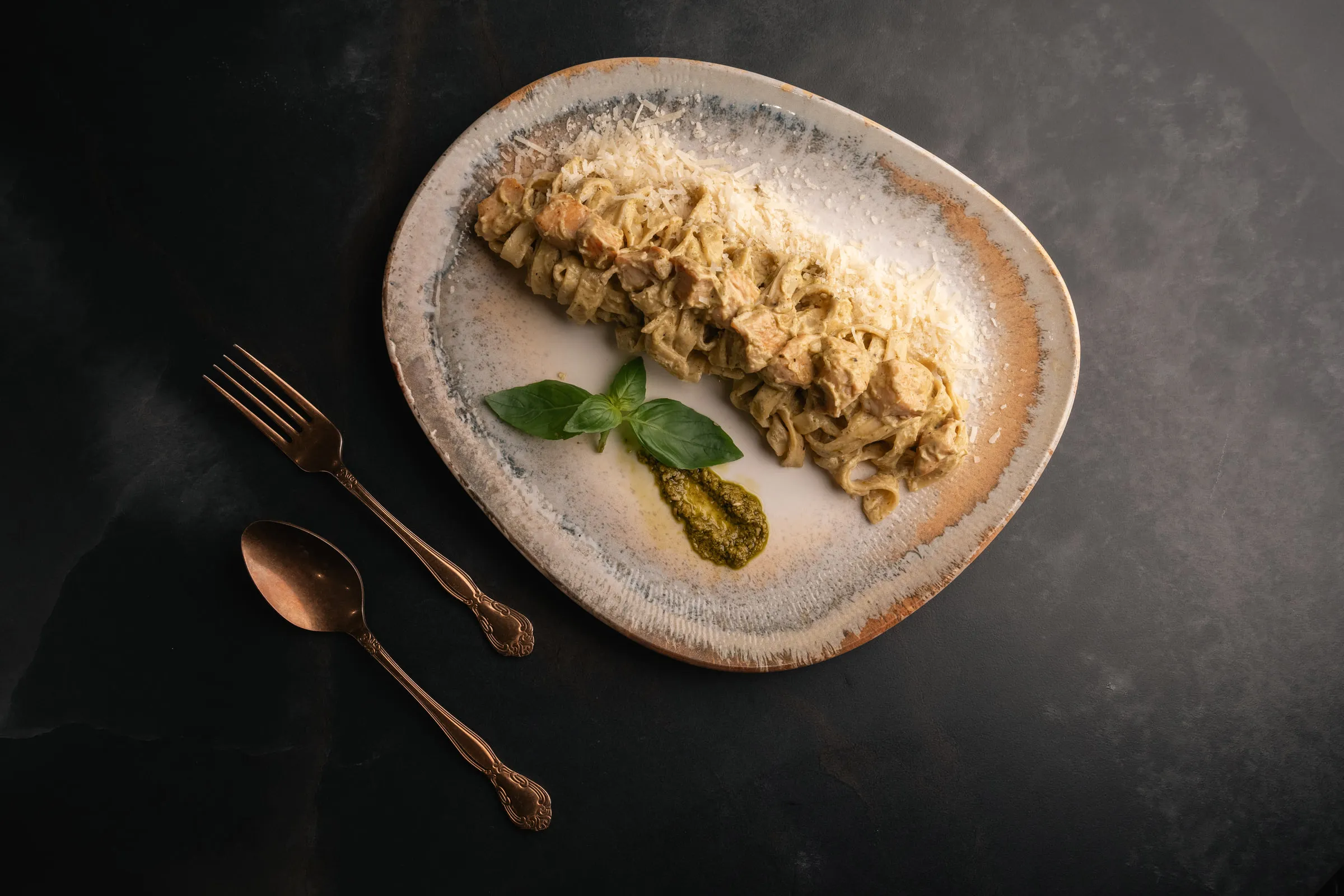 Fettuccine with chicken and pesto on a speckled plate, cutlery alongside, seen from above