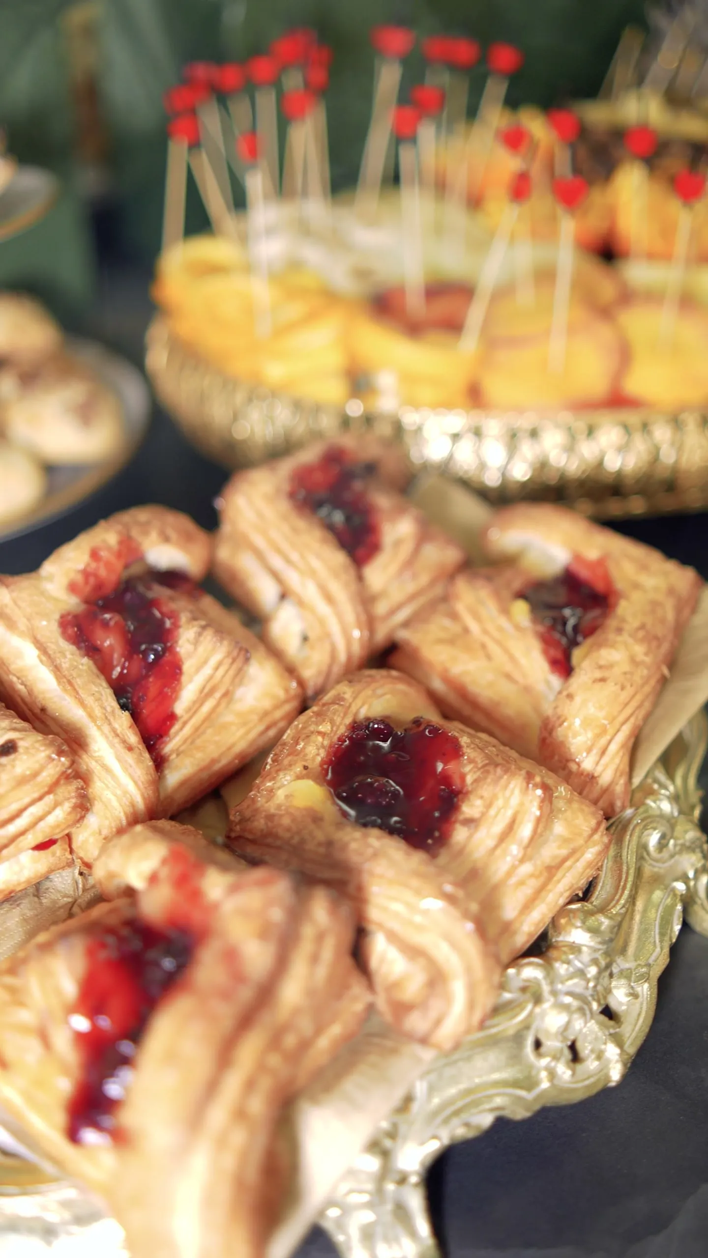 Cherry danishes on a silver tray, with a fruit tart on a gold stand behind