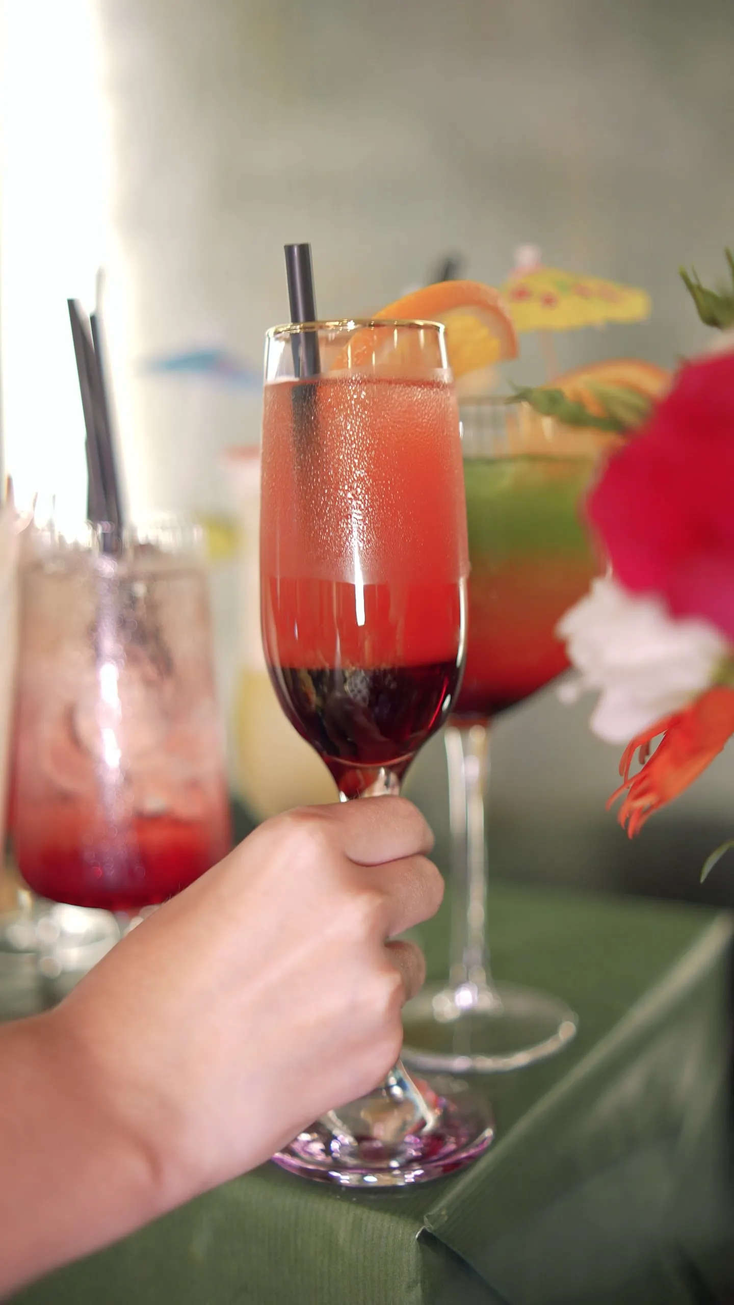 A hand holding a red mocktail in a tall flute, flowers and more drinks behind