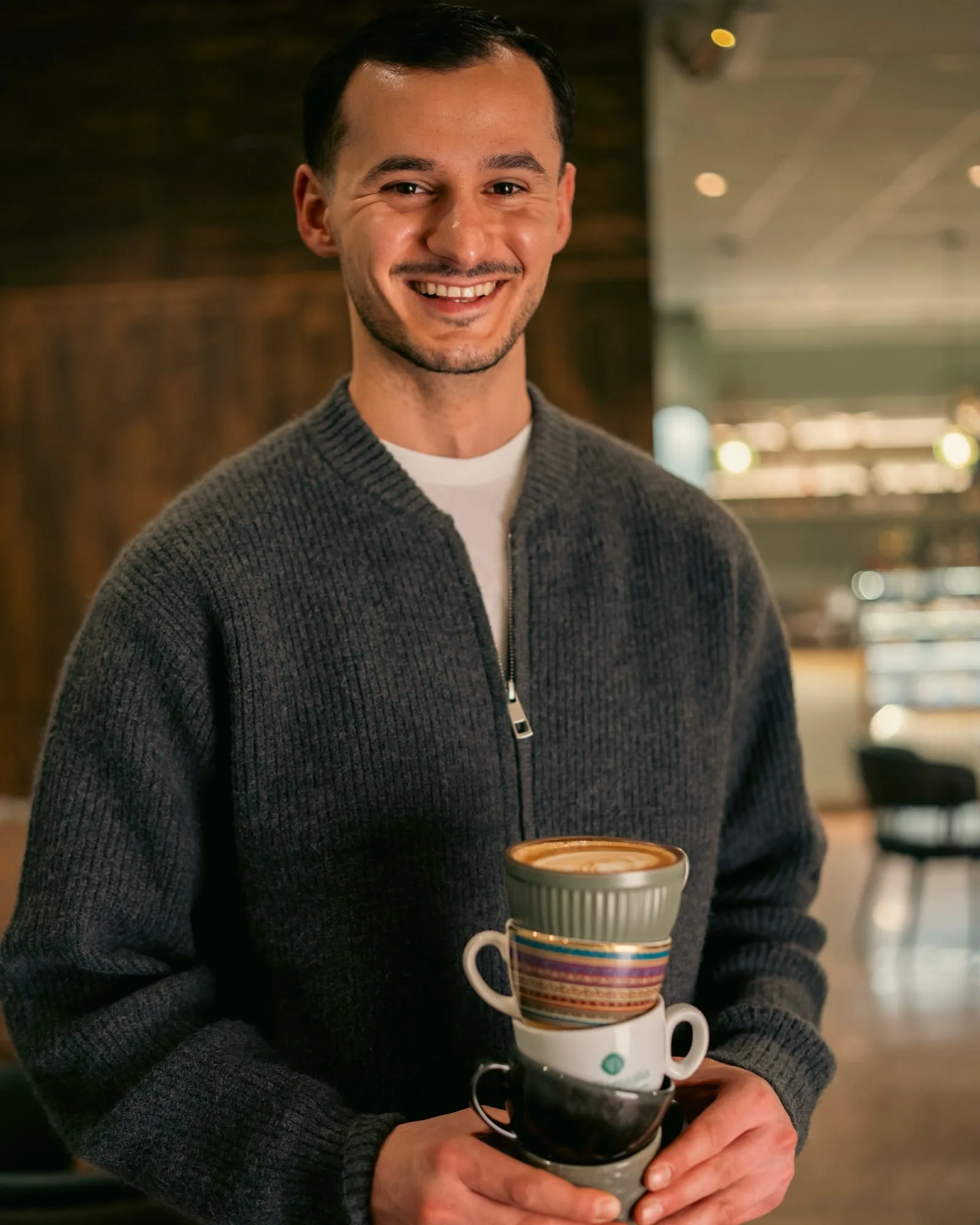 A smiling Di Famiglia barista in a grey cardigan holding a latte on a stack of colourful cups