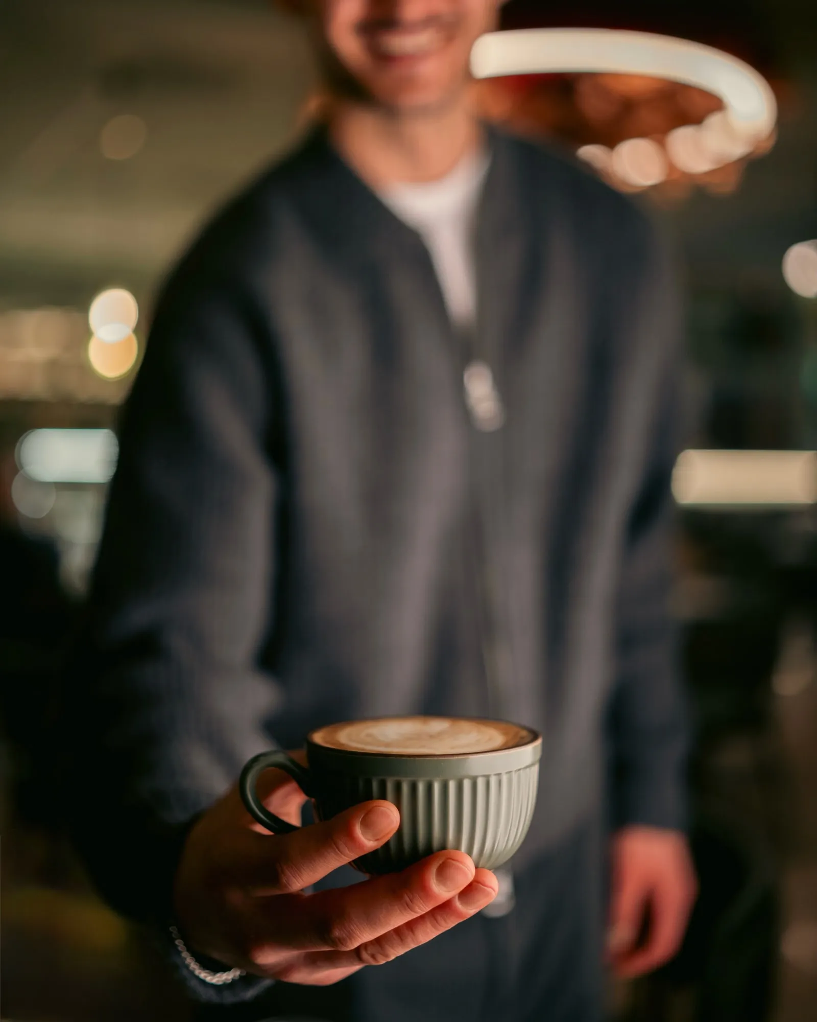 A barista holding out a latte in a ribbed grey cup, the café softly blurred behind