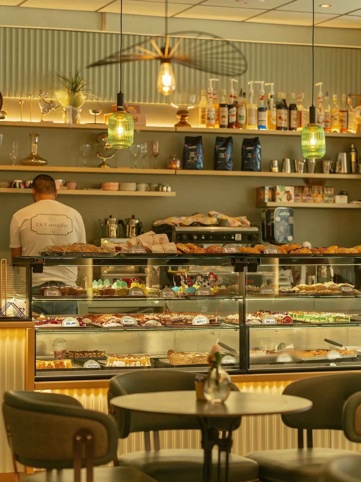 A barista at work behind the pastry vitrine, under green glass lamps and shelves of coffee and syrups